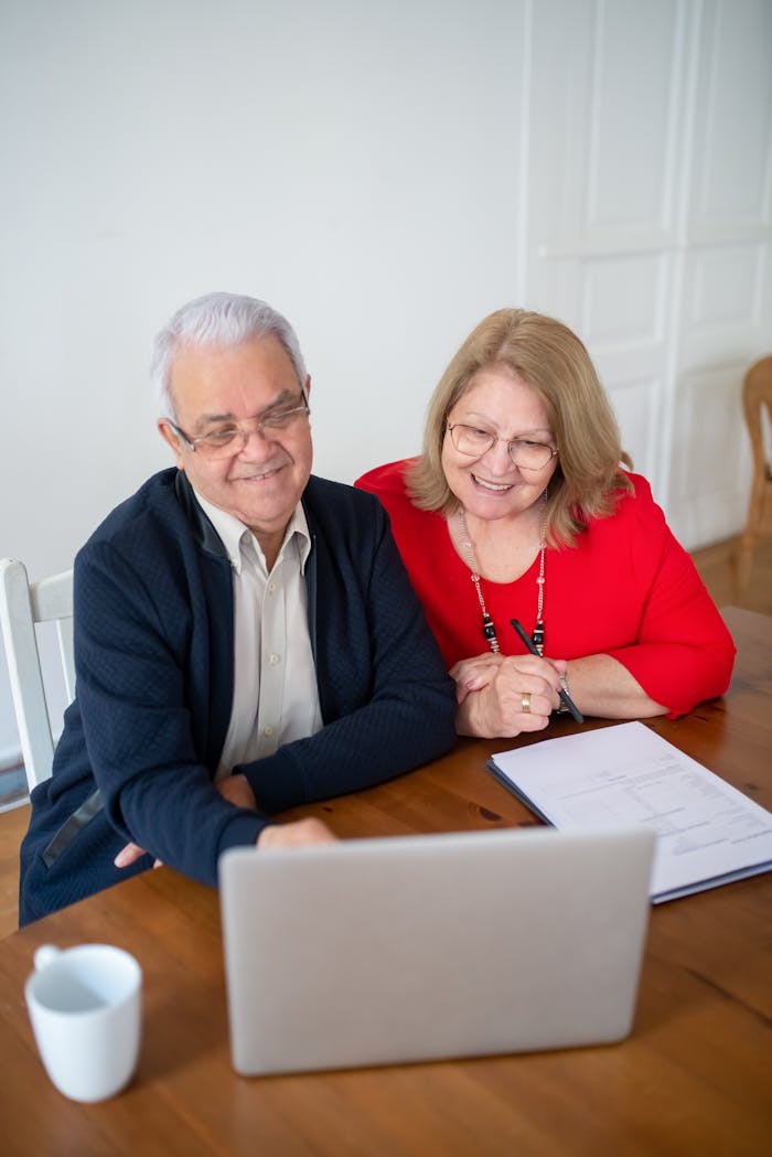 Home Senior couple sitting at a table, smiling while using a laptop indoors. Perfect for tech and lifestyle themes.