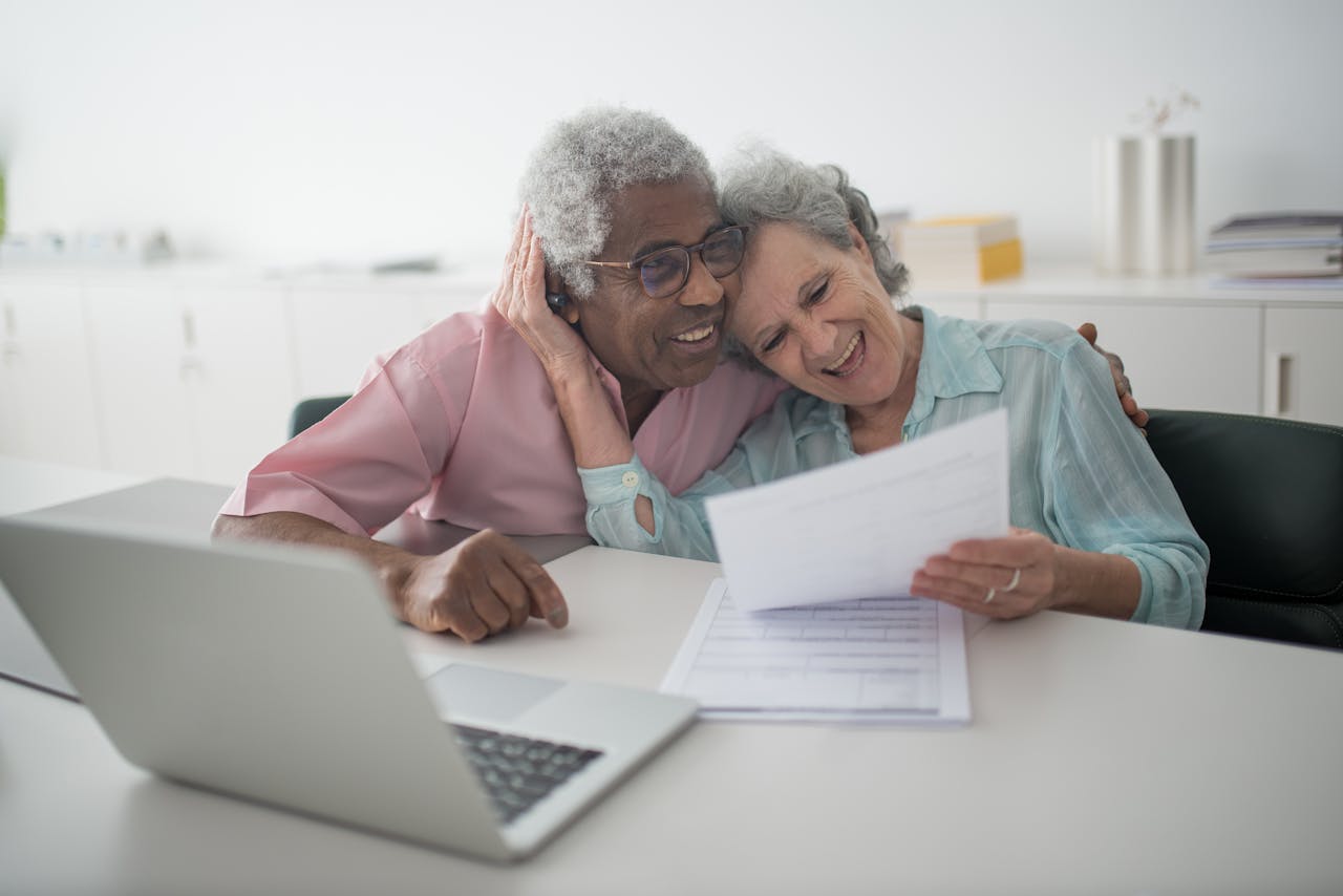 Contact A joyful senior couple smiling while reviewing documents together at a desk with a laptop.